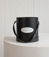 The Wild flower Signature Wild Flower black cylindrical hat box with a ribbon handle for floral arrangements.