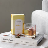 The Wild flower Libertine Blends Kapow herbal tea box with a glass of brewed tea and loose tea leaves on a book.