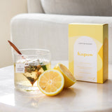 The Wild flower Kapow herbal tea box with a glass of brewed tea, a tea bag, and fresh lemon slices on a table.