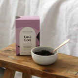 The Wild flower Libertine Blends Luxe Grey Earl Grey tea box with loose leaf tea in a ceramic bowl and copper spoon.
