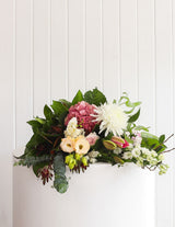 The Wild flower Pastel bouquet featuring white, pink, and nude blooms with lush greenery on a white pedestal.