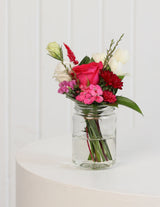 The Wild flower Petite arrangement featuring vibrant pink and white flowers in a small clear glass mason jar.