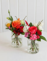 The Wild flower Petite floral arrangements featuring vibrant orange and pink roses in small glass jars on a white table.
