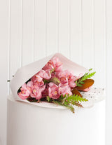 The Wild flower Cymbidium Orchid Stem bouquet wrapped in white paper with seasonal foliage on a white pedestal