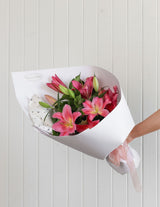 The Wild flower Lily Bouquet featuring vibrant pink lilies and green foliage wrapped in white paper with a ribbon.