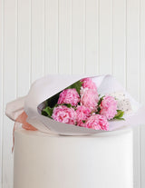 The Wild Flower Peonies Bouquet: pink peony blooms wrapped in white paper on a white pedestal