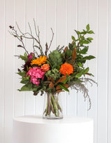 The Wild flower Florist Surprise bouquet featuring vibrant orange, pink, and deep red seasonal blooms in a glass vase.