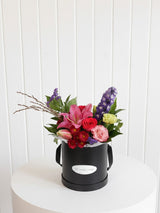 The Wild flower Wild Flower Box featuring a vibrant arrangement of seasonal pink lilies, roses, and purple delphiniums.